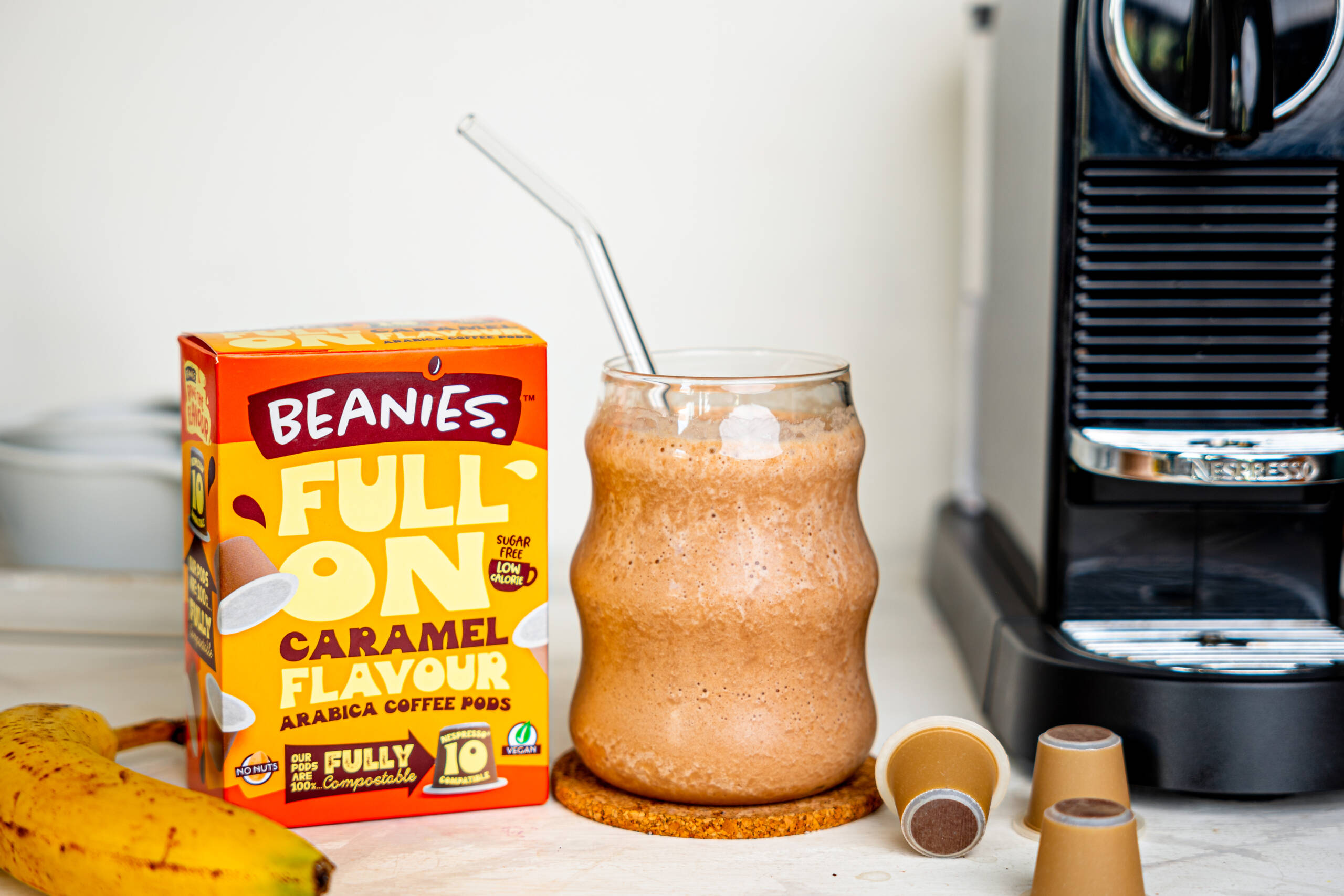Ein Glas Banana Coffee zwischen einer Nespresso-Maschine und einer Schachtel Beanies Caramel aromatisierten Kaffeepads.