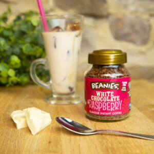 Ein frisch gebrühter Eiskaffee in einem hohen Glas hinter einer Dose Beanies White Chocolate and Raspberry Instantkaffee. Im Vordergrund des Bildes befinden sich ein Teelöffel und einige Stücke weißer Schokolade.