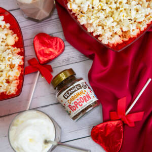Das Bild enthält in der Mitte eine Dose Beanies Cramel Popcorn Kaffee. Am Rande des Bildes befinden sich ein Kaffee mit Sahne, 2 rote, herzförmige Schalen mit Popcorn und 2 herzförmige Schokoladenlollis in roter Folie.
